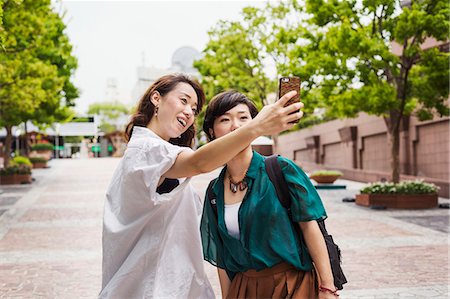 simsearch:6118-08762190,k - Two women with black hair wearing white and green shirt standing outdoors, taking selfie with mobile phone, smiling. Foto de stock - Royalty Free Premium, Número: 6118-09079530