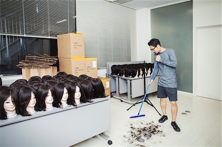 Bearded man wearing glasses standing indoors, sweeping hair on floor, large group of mannequin heads with brown wigs on tables. Stock Photo - Premium Royalty-Free, Code: 6118-09079643