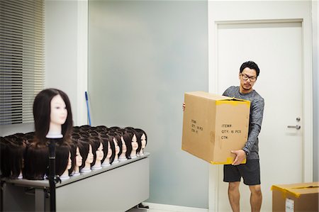 simsearch:6118-09079251,k - Bearded man wearing glasses standing indoors, carrying cardboard box, large group of mannequin heads with brown wigs on table. Foto de stock - Sin royalties Premium, Código: 6118-09079640