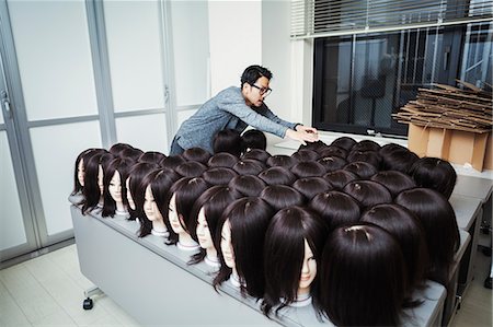 simsearch:6118-09079251,k - Bearded man wearing glasses standing indoors, arranging mannequin heads with brown wigs. Foto de stock - Sin royalties Premium, Código: 6118-09079644