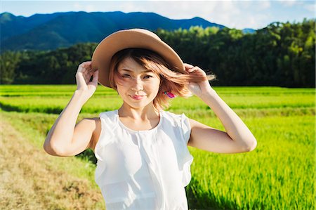 A young woman holding a straw hat on her head, hair blowing in the wind, in open space by rice paddy fields. Stock Photo - Premium Royalty-Free, Code: 6118-09079773