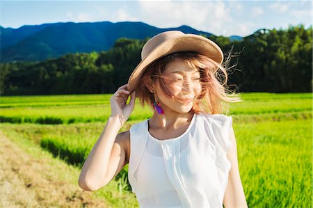 A young woman holding her hat in the wind, by rice paddy fields of green shoots, and mountain landscape. Stock Photo - Premium Royalty-Free, Code: 6118-09079774