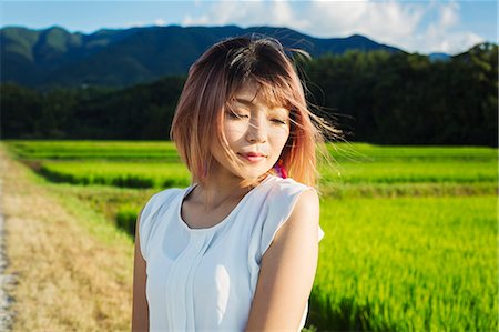 A young woman in a white shirt, hair blown in the wind, standing in open space by rice paddy fields. Stock Photo - Premium Royalty-Free, Code: 6118-09079775