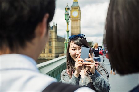 simsearch:6118-09144911,k - Smiling woman with black hair taking picture of couple with smartphone, standing on Westminster Bridge over the River Thames, London, with the Houses of Parliament and Big Ben in the background. Stock Photo - Premium Royalty-Free, Code: 6118-09079843