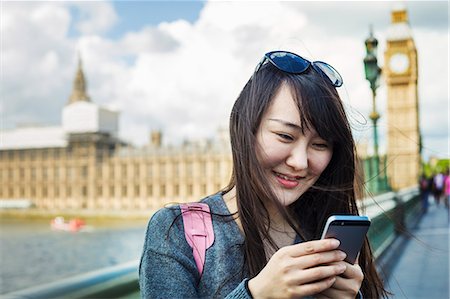 Smiling woman with black hair looking at smartphone, standing on Westminster Bridge over the River Thames, London, with the Houses of Parliament and Big Ben in the background. Stock Photo - Premium Royalty-Free, Code: 6118-09079844
