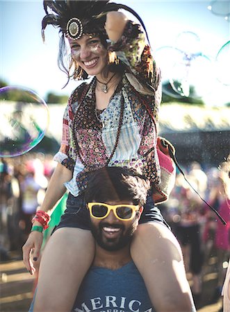 Revellers at a summer music festival young man wearing yellow sunglasses carrying woman wearing feather headdress on his shoulders. Stock Photo - Premium Royalty-Free, Code: 6118-09018292
