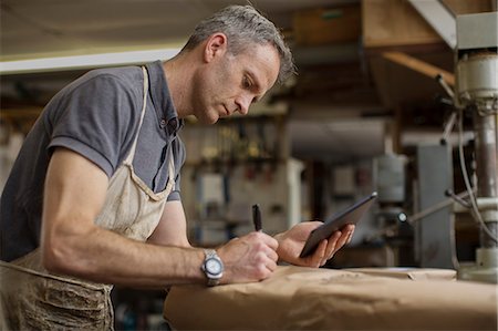 surrey - A man in a furniture restoration workshop using a digital tablet, writing on a packet wrapped in brown packaging paper. Foto de stock - Sin royalties Premium, Código: 6118-09018357