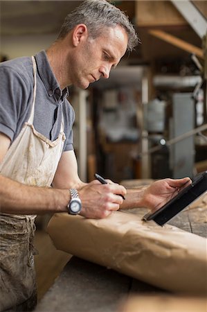 A man in a furniture restoration workshop using a digital tablet, writing on a packet wrapped in brown packaging paper Stock Photo - Premium Royalty-Free, Code: 6118-09018356