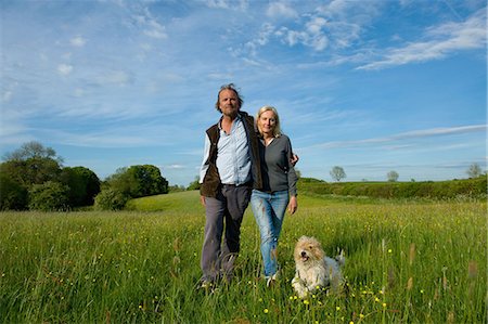 Man and woman walking arm in arm across a meadow, small dog running beside them. Stock Photo - Premium Royalty-Free, Code: 6118-09018371