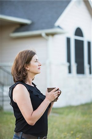 simsearch:6118-09018328,k - A woman in a sleeveless shirt holding a cup in her hands, standing outside a country house. Photographie de stock - Premium Libres de Droits, Code: 6118-09018339