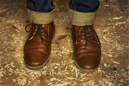 Feet and ankles of a man wearing denim jeans with turn up and traditional lace up brown leather shoes standing among wood shavings on a workshop floor. Stock Photo - Premium Royalty-Free, Code: 6118-09018601