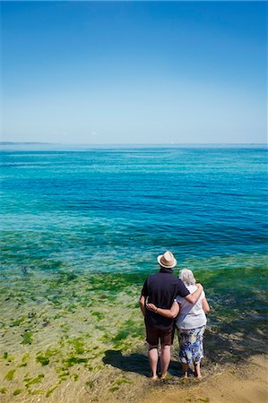 simsearch:832-03724448,k - Rear view of elderly couple standing with arms around shoulders looking out to sea across the vivid clear blue water. Foto de stock - Sin royalties Premium, Código: 6118-09018608