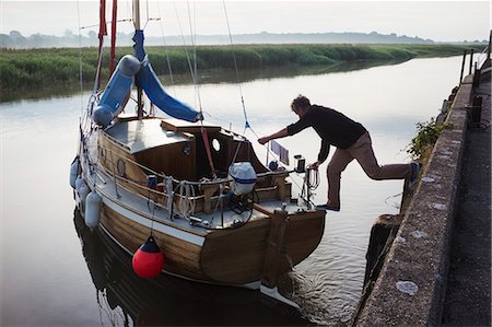 simsearch:6118-08842093,k - Man balancing between the harbour wall with one foot on a sailing boat, pushing the boat away from the mooring into the water channel. Stock Photo - Premium Royalty-Free, Code: 6118-09018633