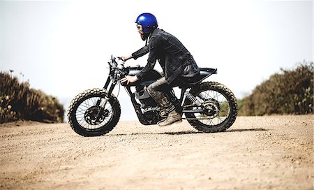 Side view of man wearing blue open face crash helmet and goggles riding cafe racer motorcycle on a dusty dirt road. Stock Photo - Premium Royalty-Free, Code: 6118-09027930