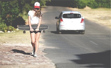 Young woman walking beside a road carrying a skateboard. Stock Photo - Premium Royalty-Free, Code: 6118-09039174