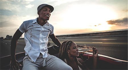 Two people riding through an urban environment in an open convertible classic car, one sitting on top of the seat back. Stock Photo - Premium Royalty-Free, Code: 6118-09039133