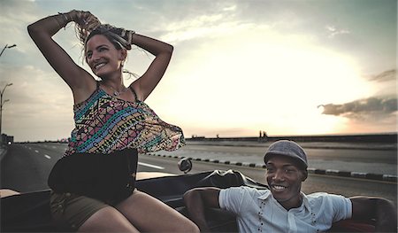 driving dusk - Two people riding through a city in an open convertible classic car, one sitting on top of the seat back. Stock Photo - Premium Royalty-Free, Code: 6118-09039134