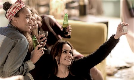 simsearch:6118-09039245,k - Four young women sitting on a sofa, laughing, taking a selfie, holding beer bottles. Foto de stock - Sin royalties Premium, Código: 6118-09039243