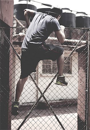 simsearch:6118-09039292,k - Young man climbing over a chain link fence in an urban environment. Foto de stock - Sin royalties Premium, Código: 6118-09039221