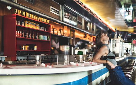 deli - Side view of woman wearing jeans and sleeveless top leaning against a bar counter in a restaurant, looking up. Stock Photo - Premium Royalty-Free, Code: 6118-09039346