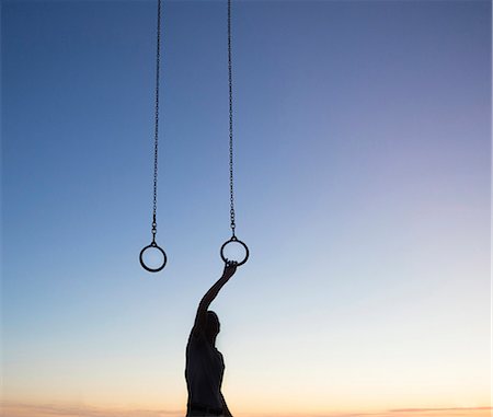 Silhouette of man standing outdoors at sunset, arm raised, holding onto gymnastic rings. Stock Photo - Premium Royalty-Free, Code: 6118-09144785
