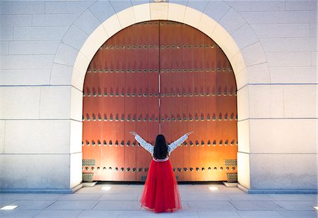 Rear view of woman wearing long red skirt standing with her arms raised in front of tall wooden gate. Stock Photo - Premium Royalty-Free, Code: 6118-09144878