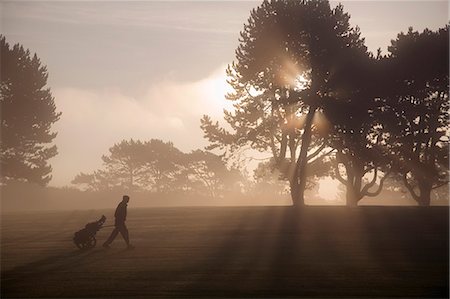 Side vie of man walking past trees across golf course at twilight, pulling golf trolley. Stock Photo - Premium Royalty-Free, Code: 6118-09144991