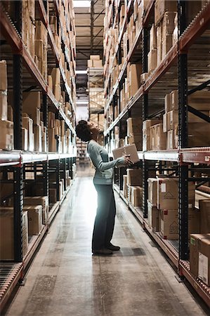 simsearch:6118-09174228,k - African American female warehouse worker checking inventory and carrying a box in an aisle of products stored in cardboard boxes, on large racks in a large warehouse distribution centre. Fotografie stock - Premium Royalty-Free, Codice: 6118-09147847