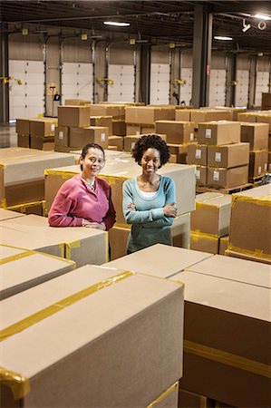 Team portrait of two African American female warehouse workers surrounded by products stored in cardboard boxes in front of loading dock doors in a large distribution warehouse. Stock Photo - Premium Royalty-Free, Code: 6118-09147845