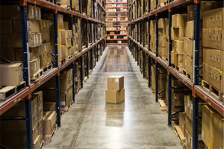 simsearch:6118-09174220,k - View down an aisle of racks holding cardboard boxes of product on pallets  in a large distribution warehouse.  Extras boxes sitting on the floor. Fotografie stock - Premium Royalty-Free, Codice: 6118-09147852