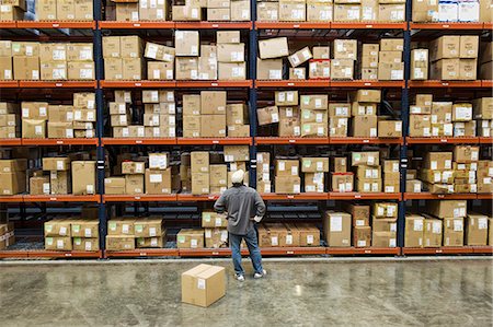 simsearch:6118-09174220,k - Warehouse worker checking inventory next to large racks of cardboard boxes holding product in a distribution warehouse. Fotografie stock - Premium Royalty-Free, Codice: 6118-09147854