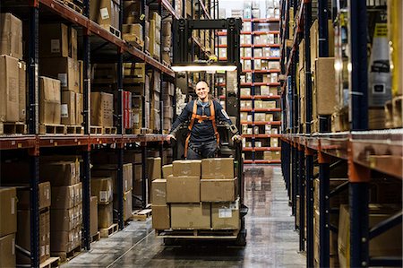 simsearch:6102-08120935,k - Warehouse worker wearing a safety harness while operating a motorized stock picker in an aisle between large racks of cardboard boxes holding product on pallets in a large distribution warehouse. Stock Photo - Premium Royalty-Free, Code: 6118-09147859