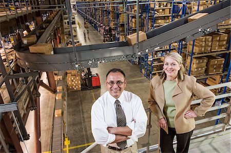 Overhead view portrait of a male Hispanic American executive in a shirt and tie and a Caucasian female executive in a jacket and slacks surrounded by large racks, forklifts and products stored in cardboard boxes  in a large distribution warehouse. Stock Photo - Premium Royalty-Free, Code: 6118-09147865