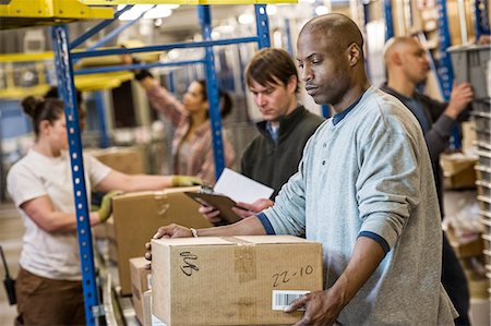 African American warehouse worker and a team of multi-ethnic workers working next to a motorized conveyor of cardboard boxes holding products in a large distribution warehouse. Stock Photo - Premium Royalty-Free, Code: 6118-09147874