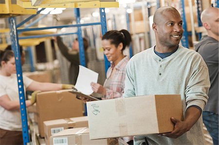 African American warehouse worker and a team of multi-ethnic workers working next to a motorized conveyor of cardboard boxes holding products in a large distribution warehouse. Stock Photo - Premium Royalty-Free, Code: 6118-09147875