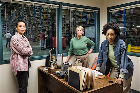 Team portrait of two African American female warehouse workers and one female Caucasian warehouse  worker in an office in the middle of a large distribution warehouse full of racks of products stored in cardboard boxes on pallets. Stock Photo - Premium Royalty-Free, Code: 6118-09147879