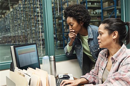 Team of two African American female warehouse workers working on a computer in an office in the middle of a large distribution warehouse full of racks of products stored in cardboard boxes on pallets. Stock Photo - Premium Royalty-Free, Code: 6118-09147880
