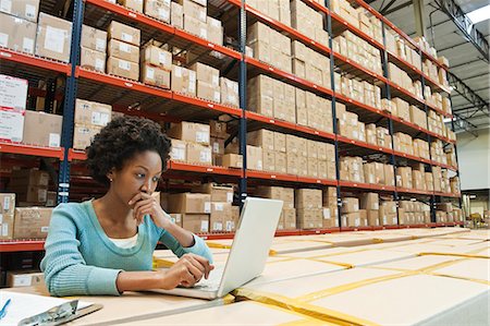 African American female warehouse worker working on inventory on a lap top computer in a large distribution warehouse of products stored in cardboard boxes and on large racks. Stock Photo - Premium Royalty-Free, Code: 6118-09147892