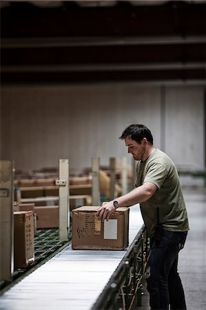 Caucasian warehouse worker in a large distribution warehouse, showing products stored in cardboard boxes, and moving on a motorized conveyor system. Stock Photo - Premium Royalty-Free, Code: 6118-09147828