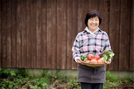simsearch:6118-08947538,k - Woman with black hair wearing checkered shirt standing in a garden, holding basket with fresh vegetables, smiling at camera. Stock Photo - Premium Royalty-Free, Code: 6118-09147915