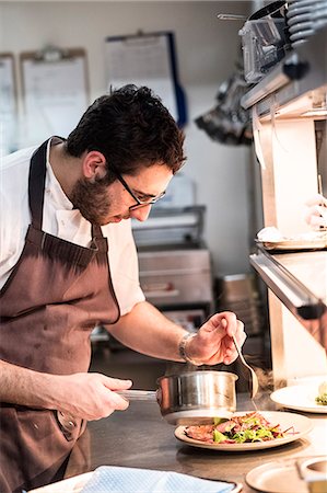 simsearch:6118-09148148,k - Bearded chef wearing glasses and apron standing in commercial kitchen at counter, plating food. Stockbilder - Premium RF Lizenzfrei, Bildnummer: 6118-09148152