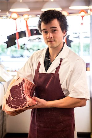 simsearch:6118-08729219,k - Man with brown curly hair wearing apron standing in butcher shop, holding beef forerib, smiling at camera. Foto de stock - Sin royalties Premium, Código: 6118-09148163