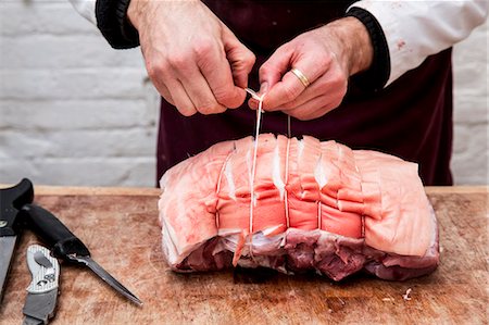 simsearch:6118-08729219,k - Close up of butcher wearing apron standing at a wooden butcher's block, preparing a rolled pork belly using string. Foto de stock - Sin royalties Premium, Código: 6118-09148167