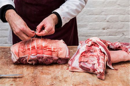 simsearch:6118-08729219,k - Close up of butcher wearing apron standing at a wooden butcher's block, preparing a rolled pork belly using string. Foto de stock - Sin royalties Premium, Código: 6118-09148168