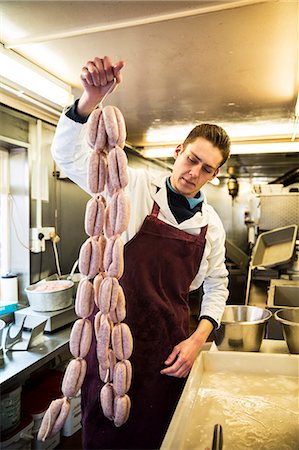 simsearch:6118-08729219,k - Man wearing apron standing in butcher shop, holding large number of freshly made sausages hanging from hooks. Foto de stock - Sin royalties Premium, Código: 6118-09148172