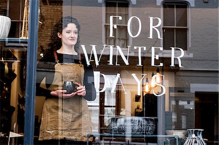 simsearch:6118-09059818,k - Woman with brown curly hair wearing apron standing behind window of pottery shop, holding mug. Stock Photo - Premium Royalty-Free, Code: 6118-09148246