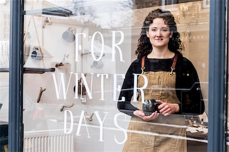 simsearch:6118-09059818,k - Woman with brown curly hair wearing apron standing behind window of pottery shop, holding mug. Stock Photo - Premium Royalty-Free, Code: 6118-09148248