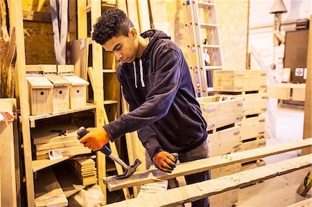 simsearch:6118-07353397,k - Young man wearing work gloves standing in warehouse, removing rusty nails from recycled wooden planks using hammer. Fotografie stock - Premium Royalty-Free, Codice: 6118-09148279