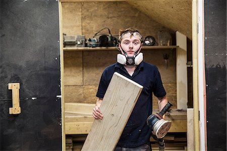 simsearch:6118-09148280,k - Young man wearing dust mask and protective goggles standing in a workshop, holding sander piece of wood. Photographie de stock - Premium Libres de Droits, Code: 6118-09148285