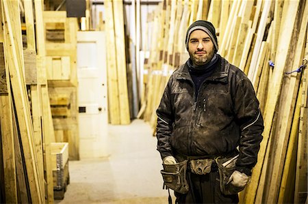 simsearch:6118-07121810,k - Bearded man wearing beanie and tool belt standing next to a stack of wooden planks in a warehouse, looking at camera. Foto de stock - Sin royalties Premium, Código: 6118-09148292